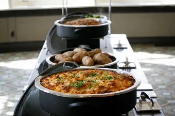 A view down the serving bar showing mac and cheese, baked potatoes, quinoa pilaf, and vegetable medley in black serving dishes.