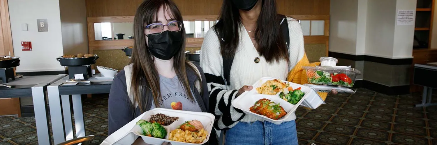 Mercy and Skylar standing in the serving line holding to-go containers full of food.