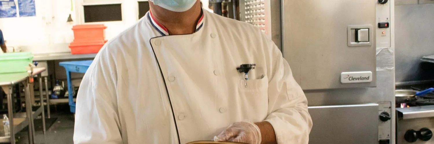Chef Mike standing the kitchen holding a to-go box of roasted chicken and carrots.