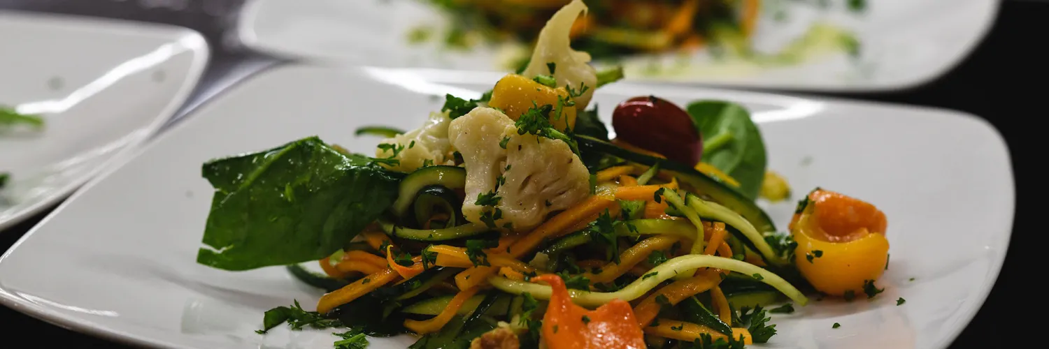 Veggie salad with tomatoes, lettuce, cauliflower, carrots, and garnish plated on a plate. Out of focus veggie salad is behind the dish in focus.